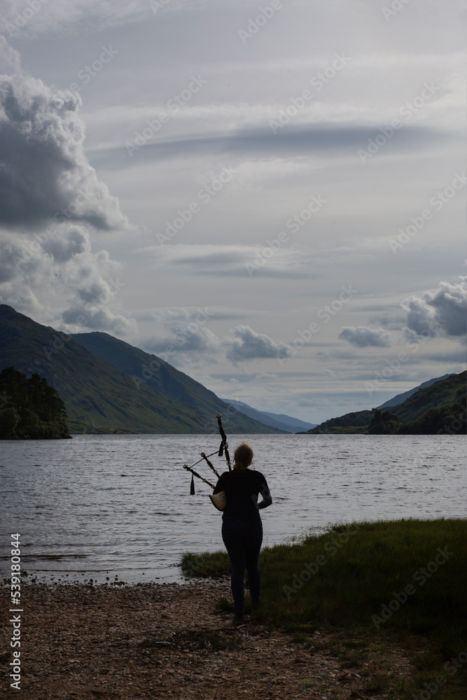 A woman plays the Scottish bagpipes on the beach at the Loch Shiel lookout, next to the Glenfinnan Monument - National Trust for Scotland