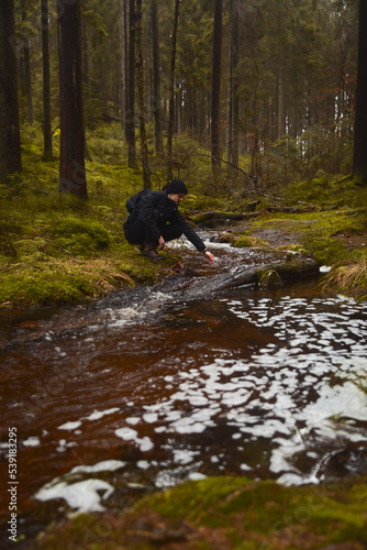 A caucasian man with a backpack sitting next to a stream in a forest touching the water.