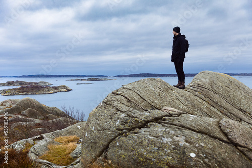 A caucasian man standing on rock by the sea watching the horizon with hiking gear on.