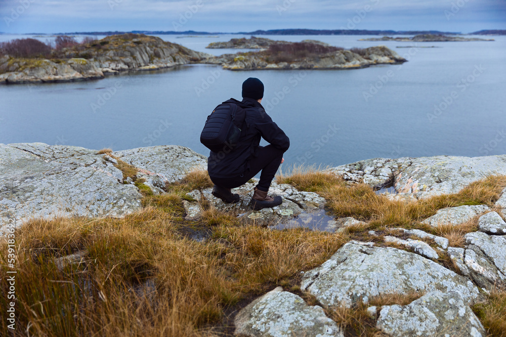 Obraz premium Hiker in black gear with a backpack crouching on a rocky cliff, looking out over a calm sea and scenic archipelago islands under a moody, overcast sky. Perfect for nature and adventure concepts.
