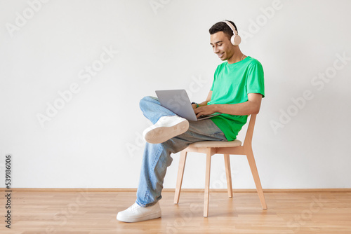 Young male freelancer working with laptop, smiling arab man sitting on chair over light wall, free space, full length © Prostock-studio