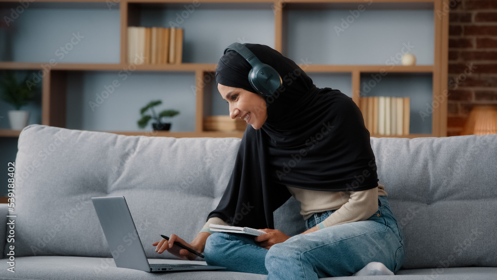 Islamic Arabian student girl Muslim woman wearing black hijab busy lady in headphones with laptop watching webinar lesson online class training write notes in notebook do homework e-learning at home Islamic Arabian student girl Muslim woman wearing black hijab busy lady in headphones with laptop watching webinar lesson online class training write notes in notebook do homework e-learning at home