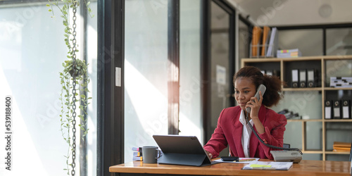 Happy african-american business woman talking on mobile at office. Young female manager consulting on phone, using laptop at working place, copy space
