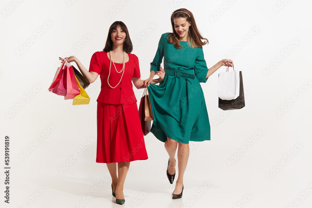 Two attractive smiling woman with color shopping bags standing on a white. Pretty young girls in retro 70s, 80s, 90s fashion style, outfits isolated in studio background. Concept of beauty, fashion