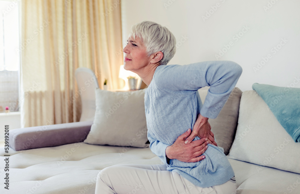Tired upset elderly woman feeling strong lower back pain after sleeping in incorrect posture on uncomfortable mattress. Senior 60s lady with closed eyes sitting on bed, holding painful lumber muscles. Tired upset elderly woman feeling strong lower back pain after sleeping in incorrect posture on uncomfortable mattress. Senior 60s lady with closed eyes sitting on bed, holding painful lumber muscles.