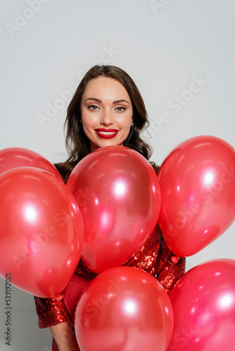 Positive woman with red lips holding festive balloons and looking at camera on grey background © LIGHTFIELD STUDIOS