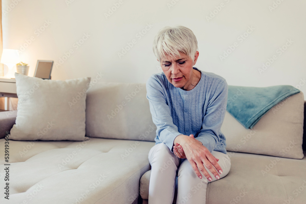 Photo of painful wrist in an elderly person. Senior woman suffering from pain in wrist at home while sitting on a gray sofa. Mature woman feeling wrist pain, injury problem, healthcare concept, sprain Photo of painful wrist in an elderly person. Senior woman suffering from pain in wrist at home while sitting on a gray sofa. Mature woman feeling wrist pain, injury problem, healthcare concept, sprain