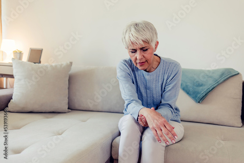 Photo of painful wrist in an elderly person. Senior woman suffering from pain in wrist at home while sitting on a gray sofa. Mature woman feeling wrist pain, injury problem, healthcare concept, sprain