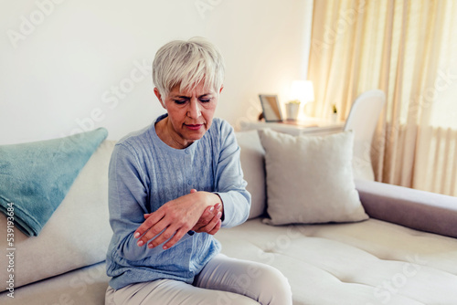 Photo of elderly lady is enduring strong ache while sitting on the sofa during the day. Senior woman at home suffering with arthritis. Upset stressed mature middle-aged woman feeling pain ache.