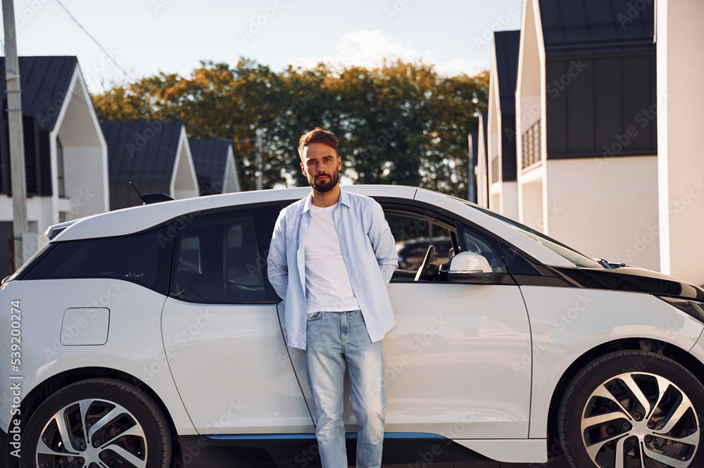 Standing and leaning on the vehicle. Posing for the camera. Young stylish man is with electric car at daytime Standing and leaning on the vehicle. Posing for the camera. Young stylish man is with electric car at daytime