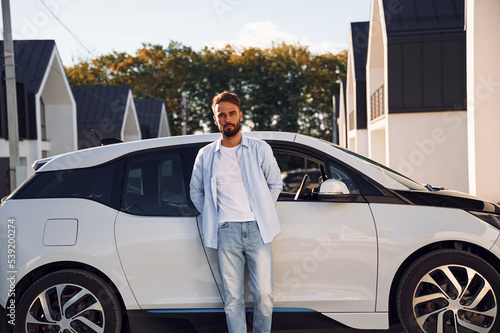 Standing and leaning on the vehicle. Posing for the camera. Young stylish man is with electric car at daytime © standret