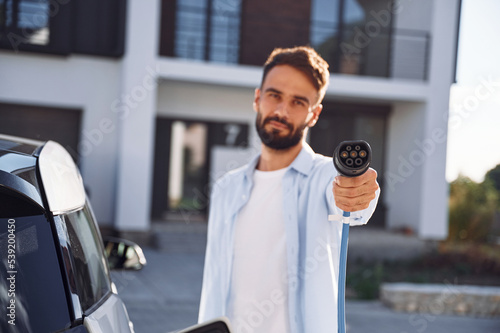 Holding and showing charging cable. Young stylish man is with electric car at daytime © standret