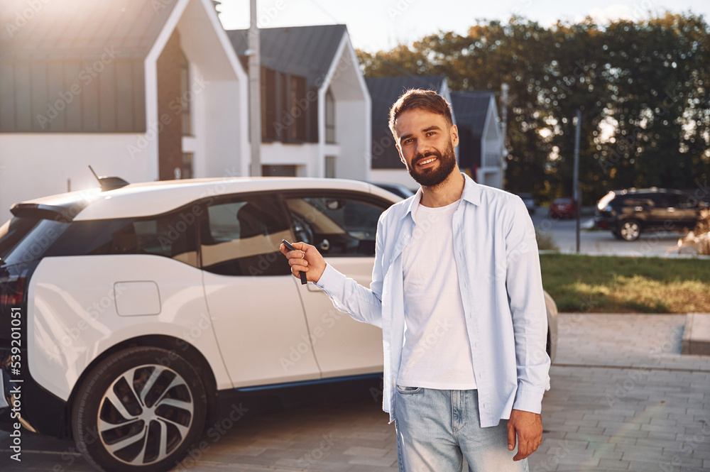 Standing and holding the keys. Young stylish man is with electric car at daytime Standing and holding the keys. Young stylish man is with electric car at daytime