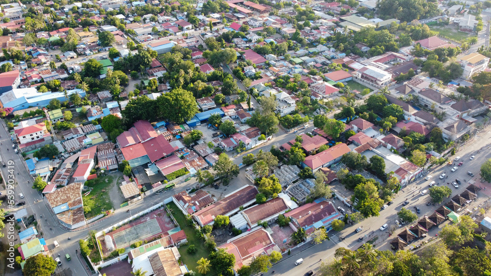 Naklejka premium Aerial drone birds eye view of bright, colorful red tin roof houses, shops and businesses surrounded by green trees within inner city capital of Dili, Timor Leste in Southeast Asia