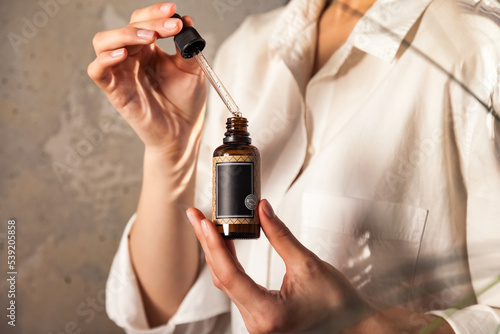 Young lady hands holding pipette and massage or cosmetics oil bottle for applying drops to skin or hair. Woman with oil at old wall background in shadow. Healthy lifestyle and self care. Copy space
