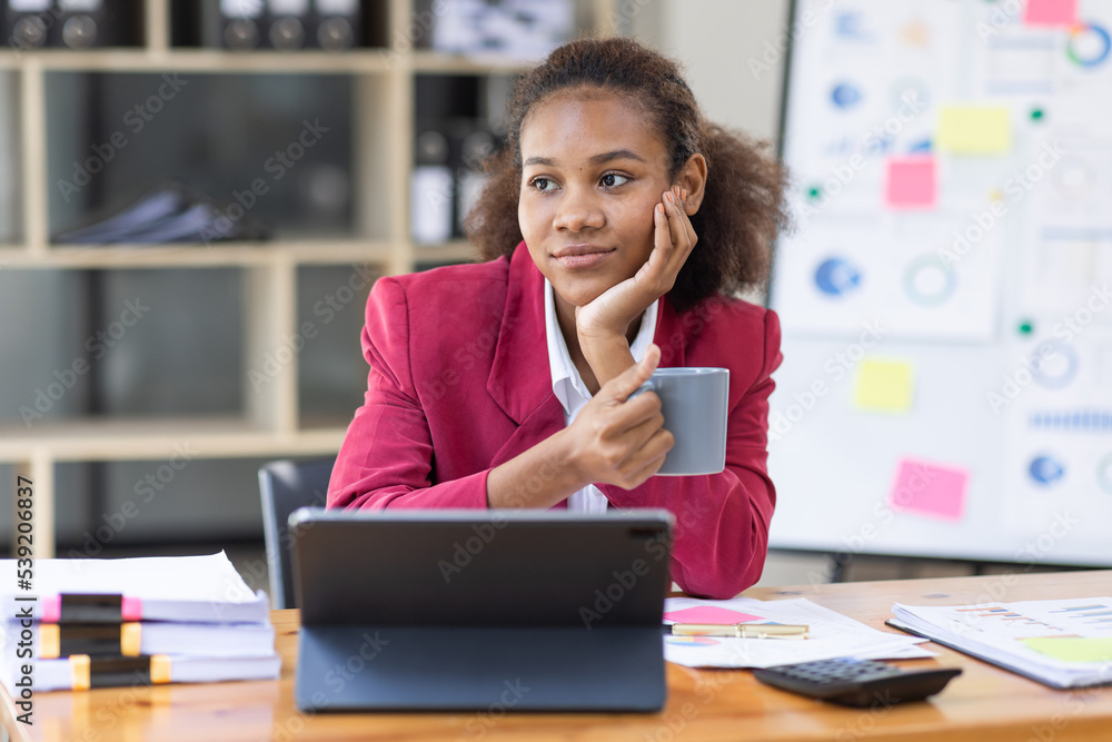 Smiling young African american female Finance Entrepreneur sitting in workplace office hoding coffee cup and working on a laptop, investment financial document report finance analysis concept. Smiling young African american female Finance Entrepreneur sitting in workplace office hoding coffee cup and working on a laptop, investment financial document report finance analysis concept.