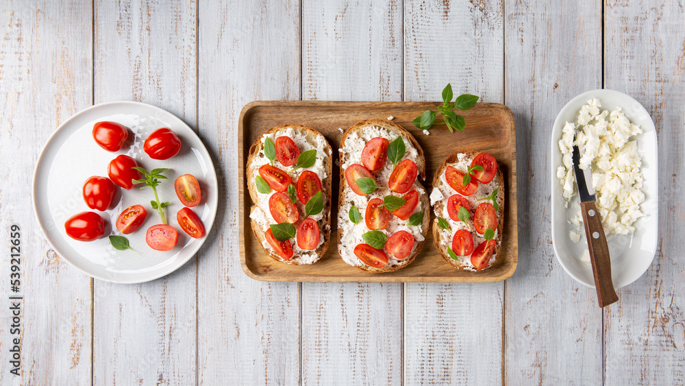 Sandwich with cottage cheese, tomatoes and basil on white wooden background. Traditional Italian bruschetta. Healthy savory feta and tomato toast. Top view. - obrazy, fototapety, plakaty 