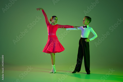 Studio shot of graceful little boy and girl dancing ballroom dance isolated over green background in neon light. Concept of art, beauty, grace, action, emotions. photo