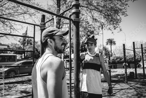 Happy, concentrated, healthy and fit young latin men resting and having a good moment between trainings in a street workout park in a sunny day (in black and white)