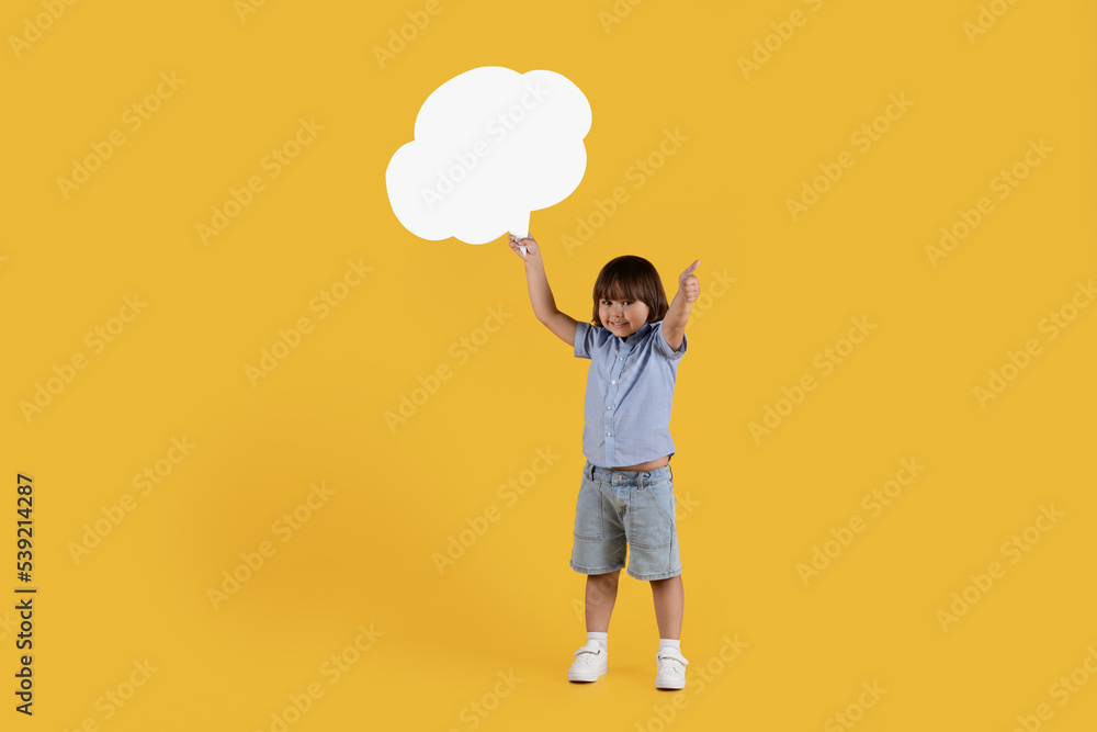 Great news. Happy little boy holding blank speech bubble and showing thumb up, smiling widely, empty space