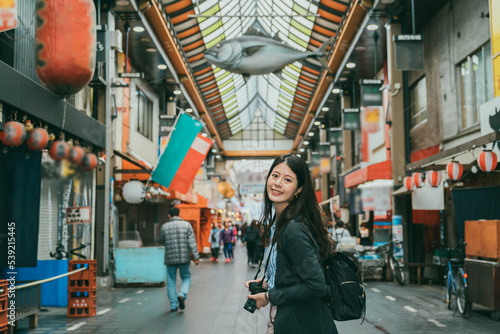 attractive Korean girl photographer turning to smile at camera in kuromon ichiba market with fish decoration hanging overhead at background in Osaka japan