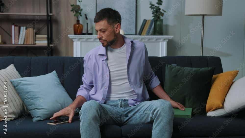 Handsome young man read book after use phone look on screen feel sad cry sitting on sofa at home. Portrait. Communication businessman chatting scrolling social media. Close up. Portrait. Slow motion Handsome young man read book after use phone look on screen feel sad cry sitting on sofa at home. Portrait. Communication businessman chatting scrolling social media. Close up. Portrait. Slow motion