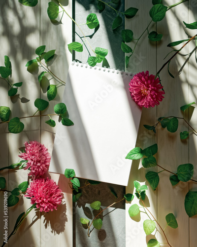 An open blank notebook, flowers, a plant with green leaves are on a white wooden background. Abstract botanical composition with copy space. The concept of taking notes, keeping a diary, to-do list.