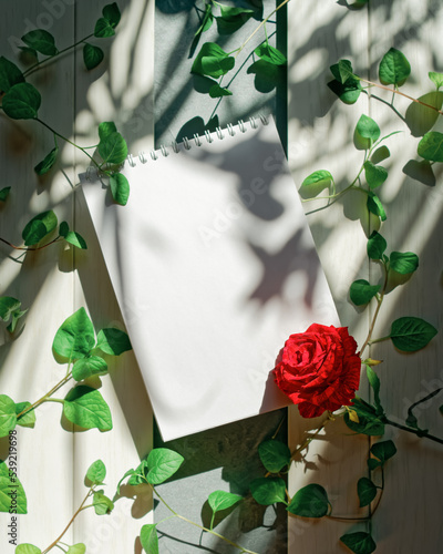 An open blank notebook, a red rose and a plant with green leaves are on a white wooden background. Romantic composition with copy space. The concept of wedding planning, taking notes, to-do list.