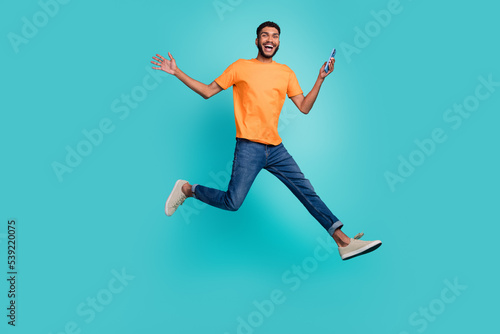 Full length photo of excited positive guy wear orange t-shirt jumping high chatting modern gadget isolated teal color background