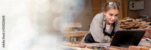 web banner Portrait of a female carpenter looking at designs on a tablet for making her furniture in a furniture factory. with many tools and wood with modern tools with copy space on left