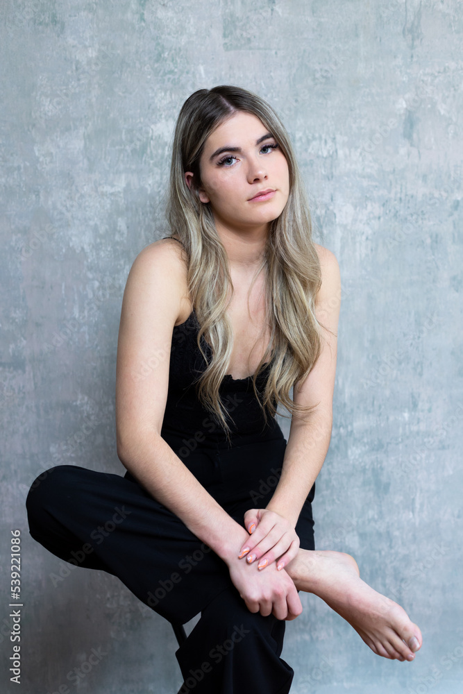 Fototapeta premium Vertical portrait of beautiful blue-eyed slim young woman with long dirty blond hair sitting on stool in black top and dark jeans in front of painted canvas background