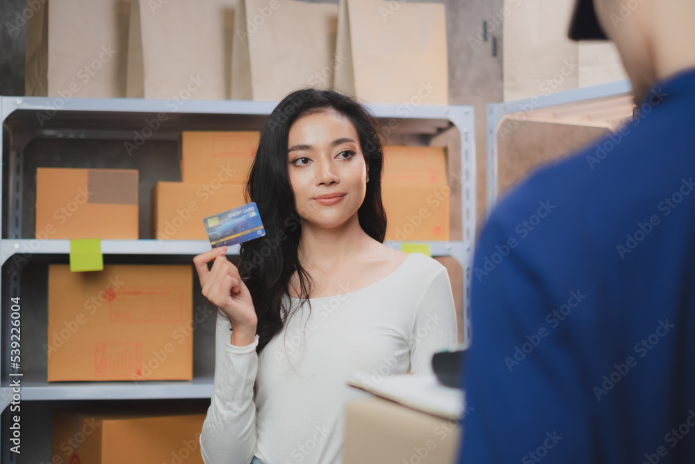 Asian female SME owners or businesswoman is holding credit cards to pay an Online order delivery. Starting a small business SME. Messenger come to pick up a box at home. Focus on Face. 
