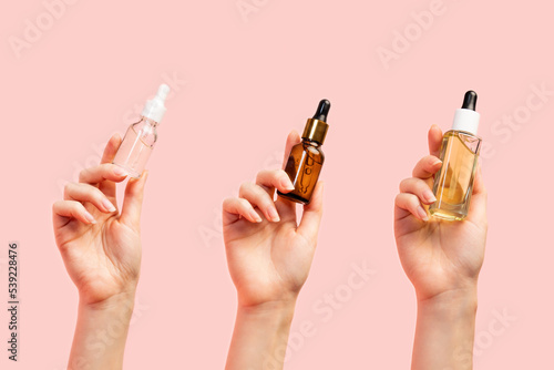 Three female hands of different nationalities hold glass multi-colored bottles with oil and serum. Pink background, close-up. Copy space. The concept of care cosmetics for any skin type photo