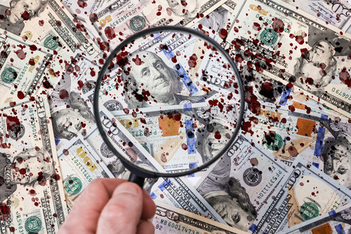 Fototapeta Naklejka Na Ścianę i Meble -  A hand with a magnifying glass against the background of scattered American dollar bills with blood drops, close-up. Flat lay. The concept of counterfeit money, bribery and illegal corruption