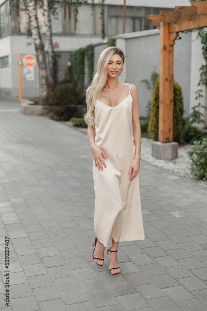 Fashion pretty smiling woman model with slender body in fashionable beige long strappy dress with shoes walks on the street