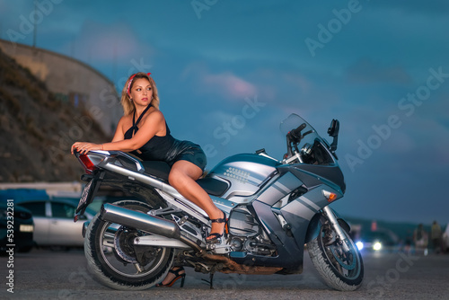 World motorcyclist day. Elegant beautiful woman confidently poses lies a motorcycle. Dusk sky at background. The concept of motorbike travel