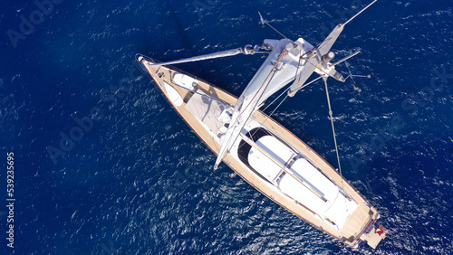 Aerial drone top down photo as seen from top of mast of beautiful sail boat with wooden deck anchored in deep blue Aegean sea