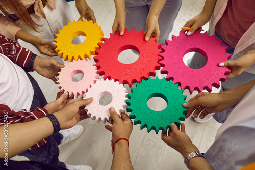 Creative young people hold colored gears of different sizes symbolizing coordinated teamwork. Top view from angle of hands of men and women standing in circle. Concept of team and teamwork.