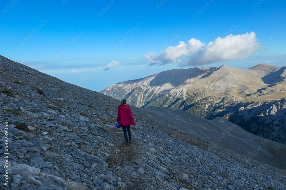Rear view of woman with climbing helmet walking on cloud covered mountain summit of Skolio Mount Olympus, Mt Olympus National Park, Macedonia, Greece, Europe. View of rocky ridges and mountain ranges