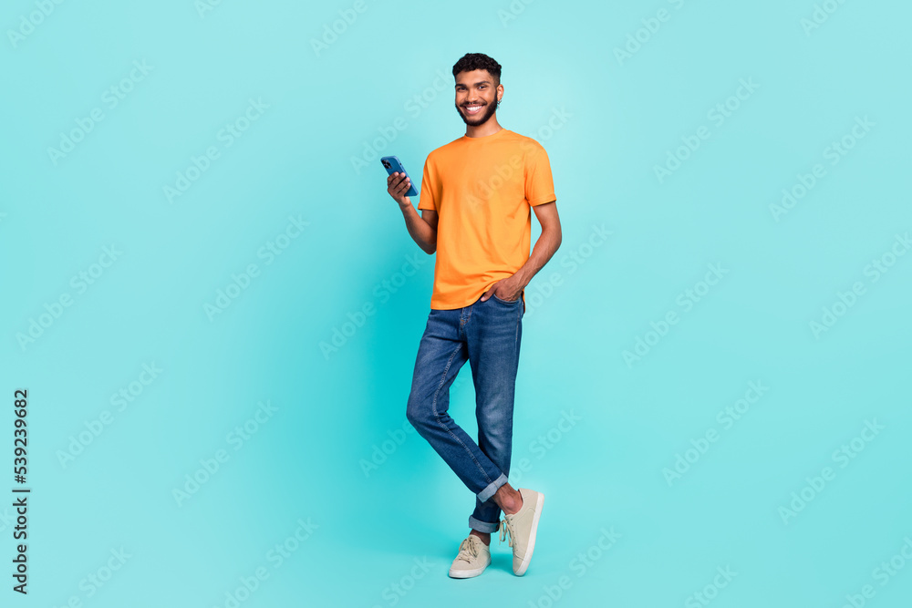 Full length photo of good funny pretty guy wear orange t-shirt communicating modern gadget empty space isolated teal color background Full length photo of good funny pretty guy wear orange t-shirt communicating modern gadget empty space isolated teal color background