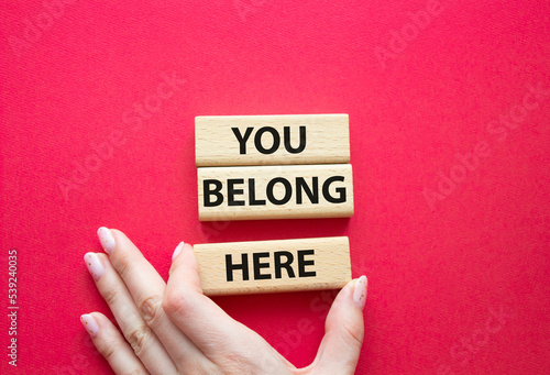 You belong here symbol. Wooden blocks with words You belong here. Beautiful red background. Businessman hand. Business and You belong here concept. Copy space.