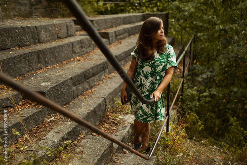 A young brunette woman with a green flower dress and books standing on the top of a hill on rocks leaning on a black fence in the forest in summer.