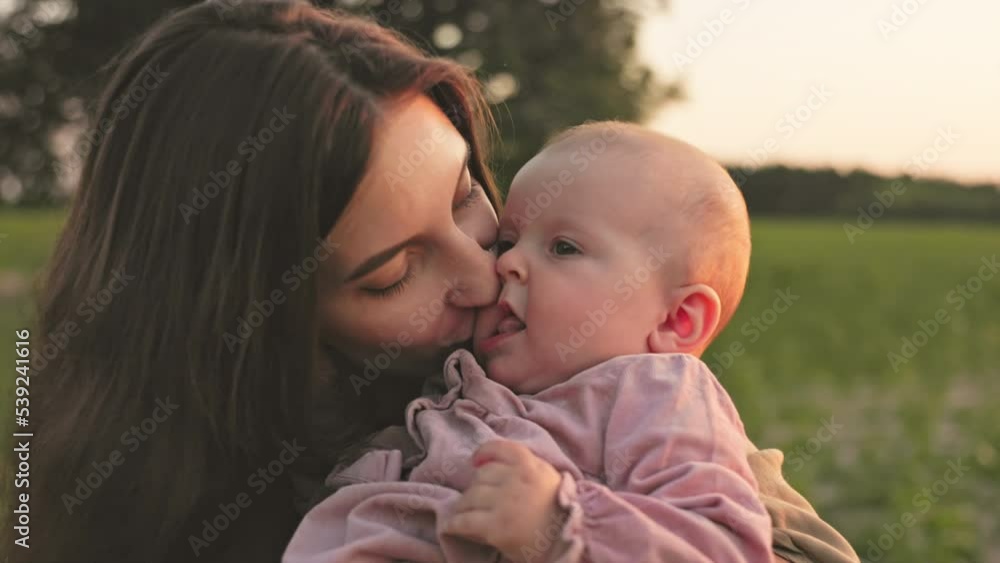 Happy baby sitting in mother's arms. Close-up shot of young good-looking woman kissing lovely daughter. Positive emotions. Horizontal view. Sunset on field in summertime. Footage of attractive Happy baby sitting in mother's arms. Close-up shot of young good-looking woman kissing lovely daughter. Positive emotions. Horizontal view. Sunset on field in summertime. Footage of attractive