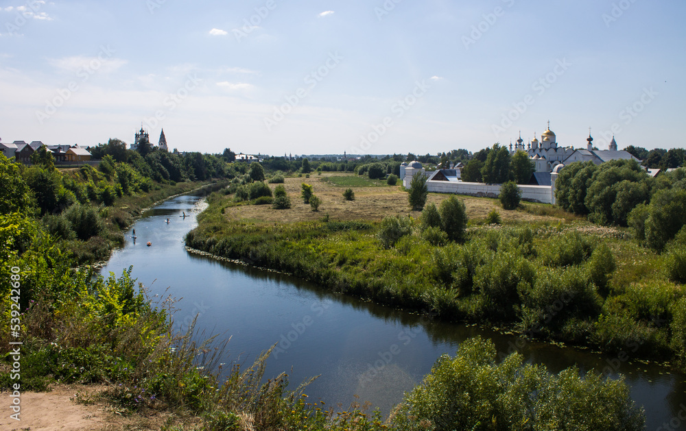 Pastoral landscape - a beautiful bend of the Kamenka river among a green meadow with grass and a white stone monastery on the shore on a sunny summer day in Suzdal, Vladmir region, Russia