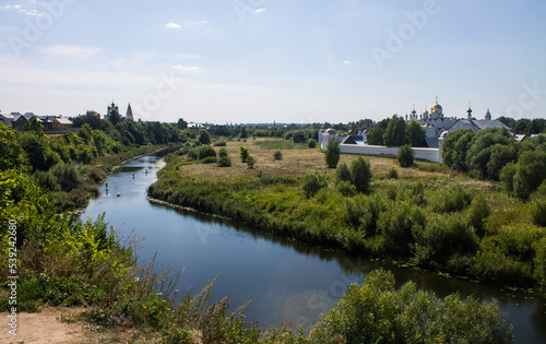 Pastoral landscape - a beautiful bend of the Kamenka river among a green meadow with grass and a white stone monastery on the shore on a sunny summer day in Suzdal, Vladmir region, Russia
