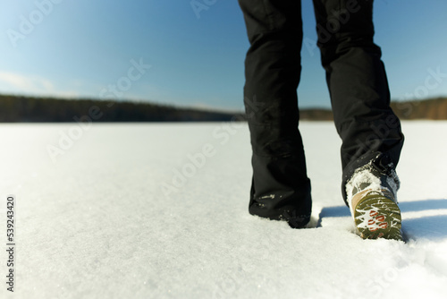 No face photo. Picture of legs in black warm clothes and green shoes walking in snow drift on white field with forest on horizon, spending sunday winter morning in rural area, far away from city