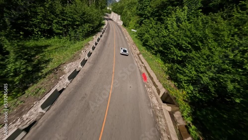 Fall from above to sport car riding on slope serpentine road. FPV drone asphalt highway level automobile fast driving up mountain peak nature scenery. Aerial view summer sunny alpine forest resort 4k