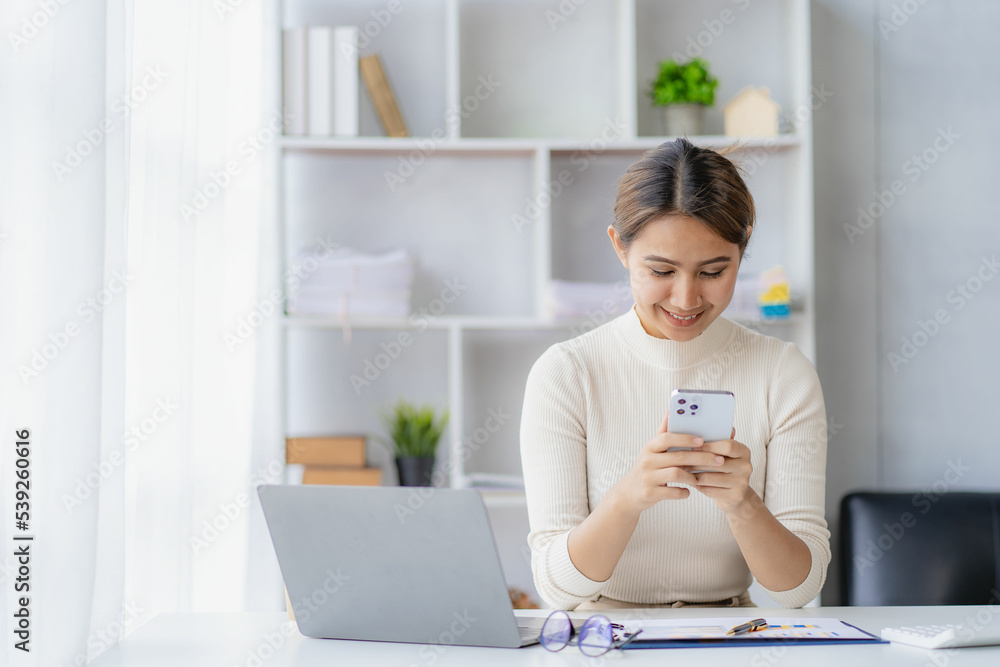 Successful Asian businesswoman smiling using a smartphone talking to a client at her desk in the office of a financial accounting concept company. Successful Asian businesswoman smiling using a smartphone talking to a client at her desk in the office of a financial accounting concept company.