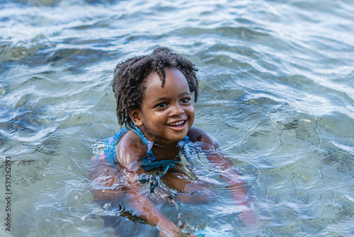 Imagen horizontal de una hermosa niña afroamericana de cabello afro muy sonriente sola jugando en el mar en un día de verano. 
