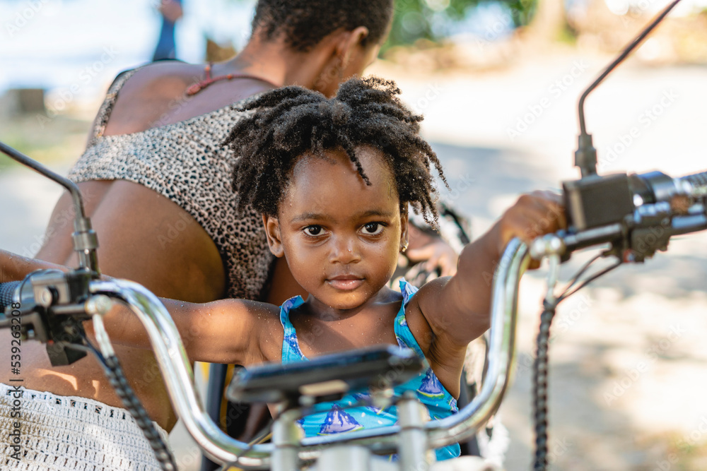 Obraz premium Imagen horizontal de una pequeña niña afroamericana mirando a cámara mientras se sostiene del manubrio de una motocicleta. 